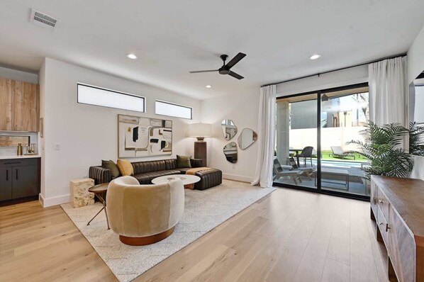 A modern living room with a sofa, armchair, and coffee table, a ceiling fan, and a view of the outdoors through large glass doors. - East Austin 5BR w Private Pool, Rooftop & Views! (Austin)