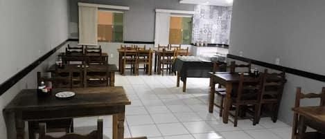 A dining area with multiple wooden tables and chairs, a tiled floor, and a view of a kitchen area in the background.