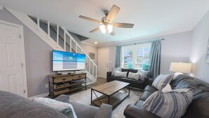 A living room with a TV, sofa, coffee table, and staircase. - Sand Dollar 5 – Your Bright & Breezy East Beach Getaway!  Free Daily Activities! (Gulf Shores)