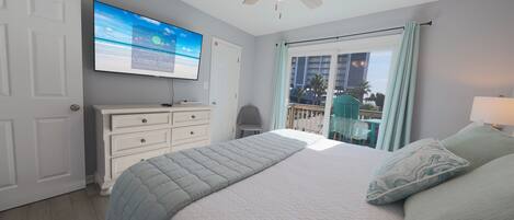 A bedroom with a bed, a ceiling fan, a TV on the wall, and a view of the outdoors.