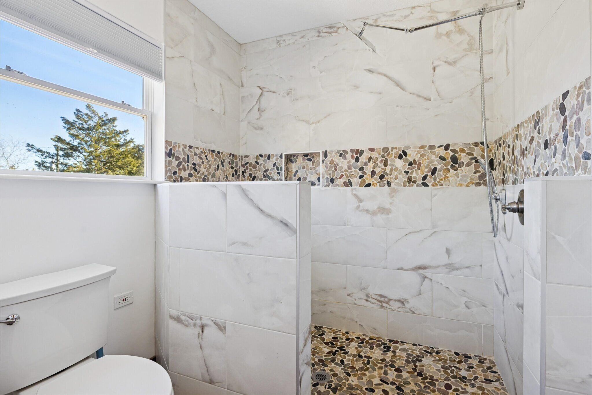 A bathroom with a shower, marble tiles, a pebbled backsplash, and a window.