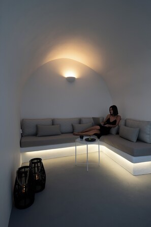 A modern living room with a curved sofa, a small coffee table, and two black candle holders. - ORANA Residence Santorini (Thira - Santorini)