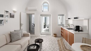 A modern interior with a sofa, a small table, a kitchen area, and a view through an open door. - ORANA Residence Santorini (Thira - Santorini)