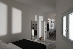 A bedroom with a bed, a small desk, a chair, and a kitchenette in the background. - ORANA Residence Santorini (Thira - Santorini)