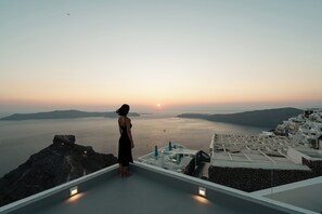 A woman standing on a rooftop overlooking a coastal landscape with a sunset. - ORANA Residence Santorini (Thira - Santorini)