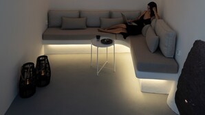 A modern, minimalist interior with a curved white wall, a sofa, a small table, and two black chairs. - ORANA Residence Santorini (Thira - Santorini)