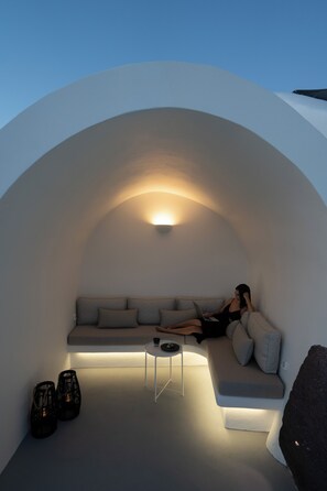 A modern, minimalist interior with a curved white wall, a sofa, a small table, and two black chairs. - ORANA Residence Santorini (Thira - Santorini)