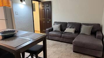A compact living space with a kitchenette, a dark brown leather sofa with cushions, a dining table with a bowl, and a tiled floor.