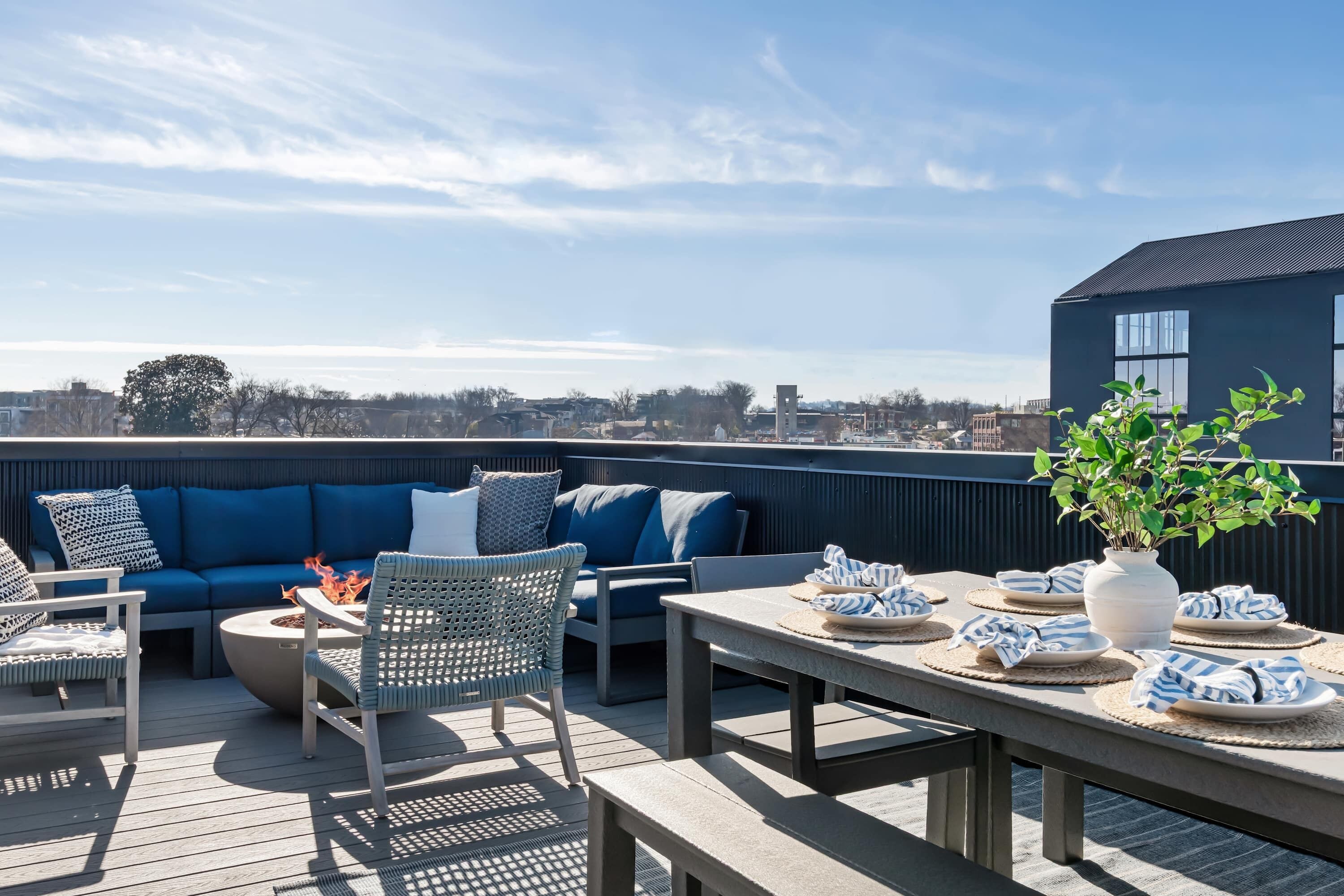 Eine Dachterrasse mit blauer Couch, Korbstühlen, einer Feuerstelle und einem Esstisch, gedeckt mit Tellern und Servietten.