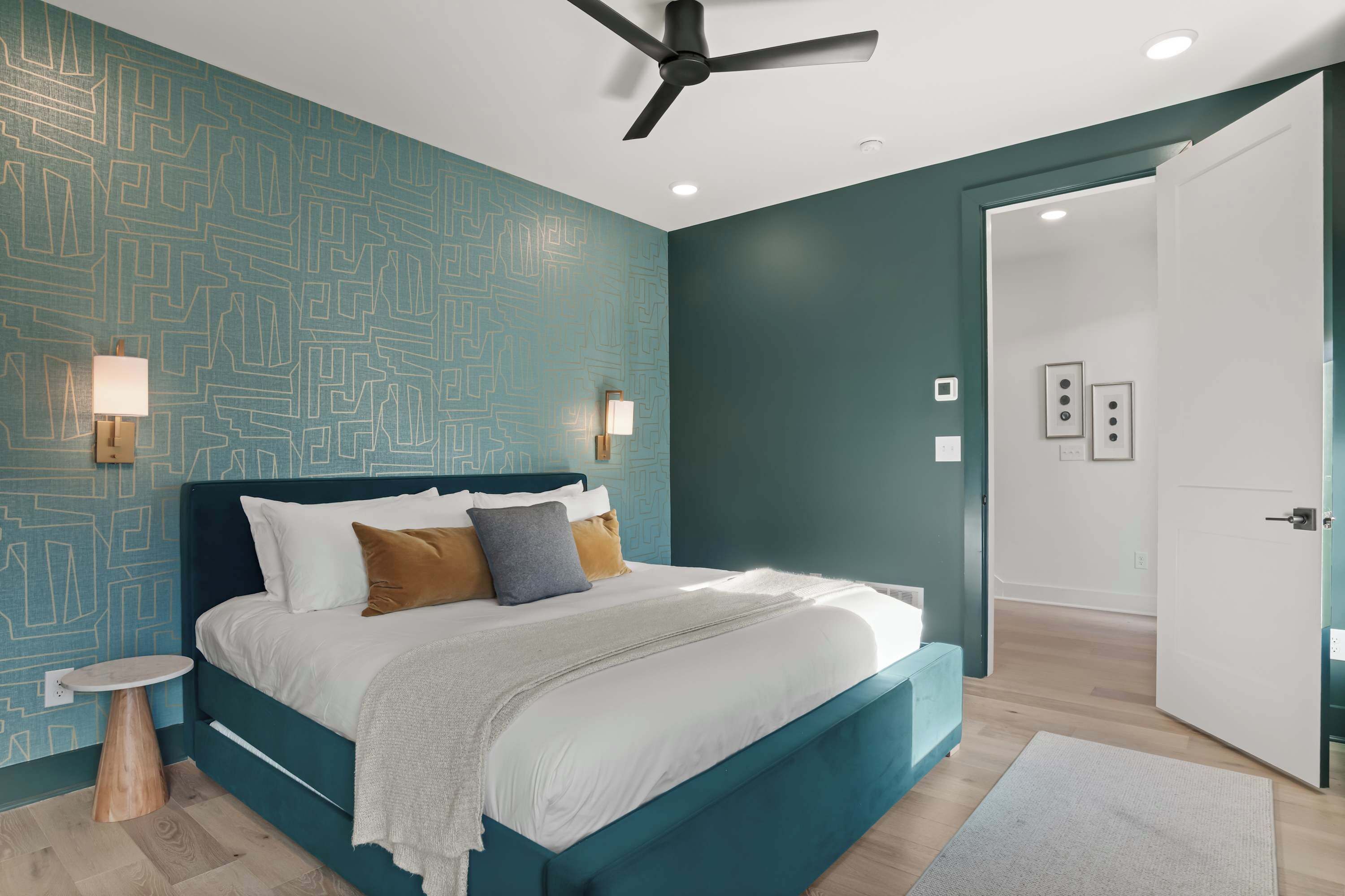 A modern bedroom with a teal-colored wall, a large bed with white bedding, a round side table, and a ceiling fan.