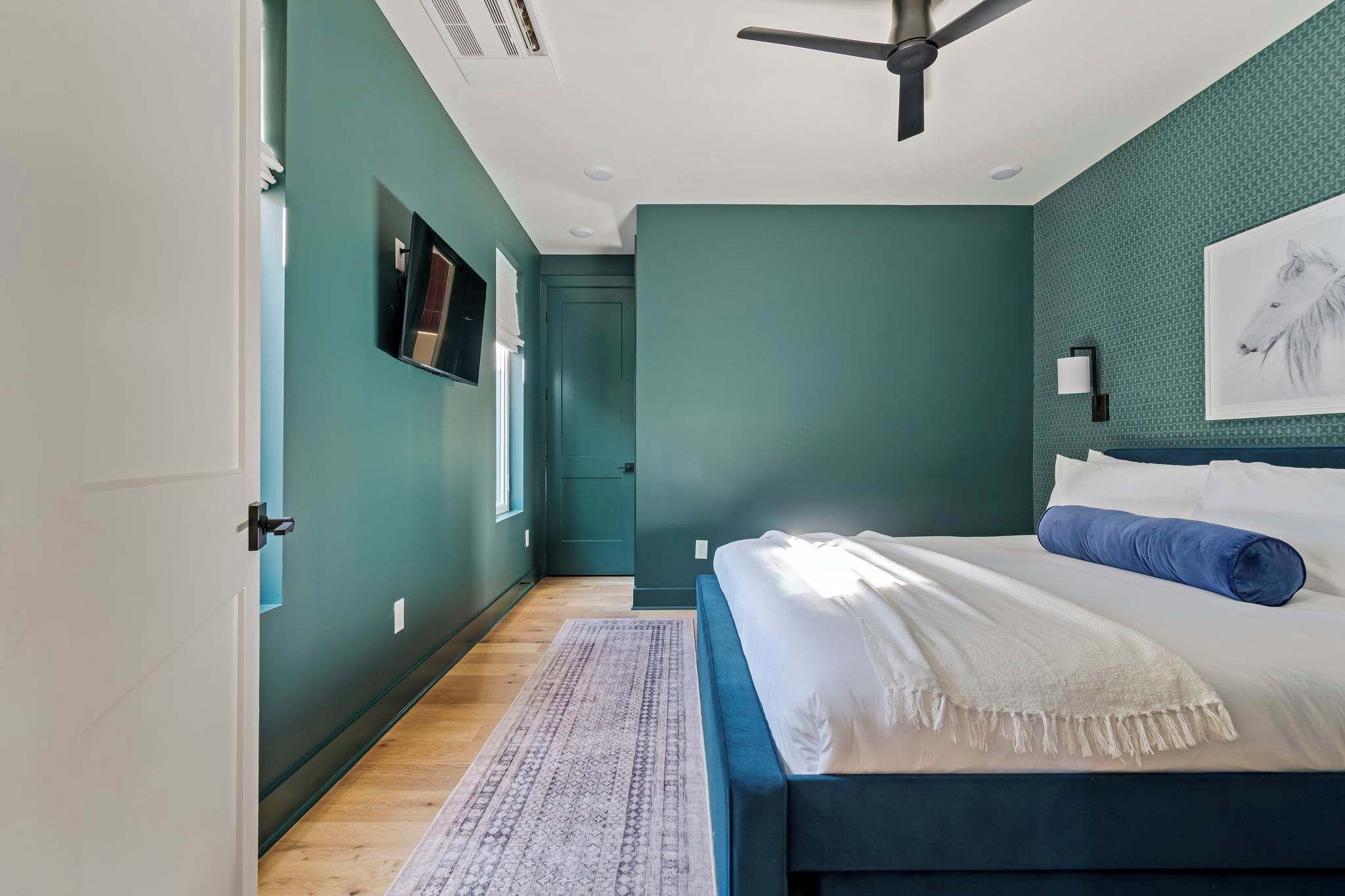 Une chambre à coucher comprenant un lit, un ventilateur de plafond, un téléviseur fixé au mur et une porte donnant sur une autre pièce.