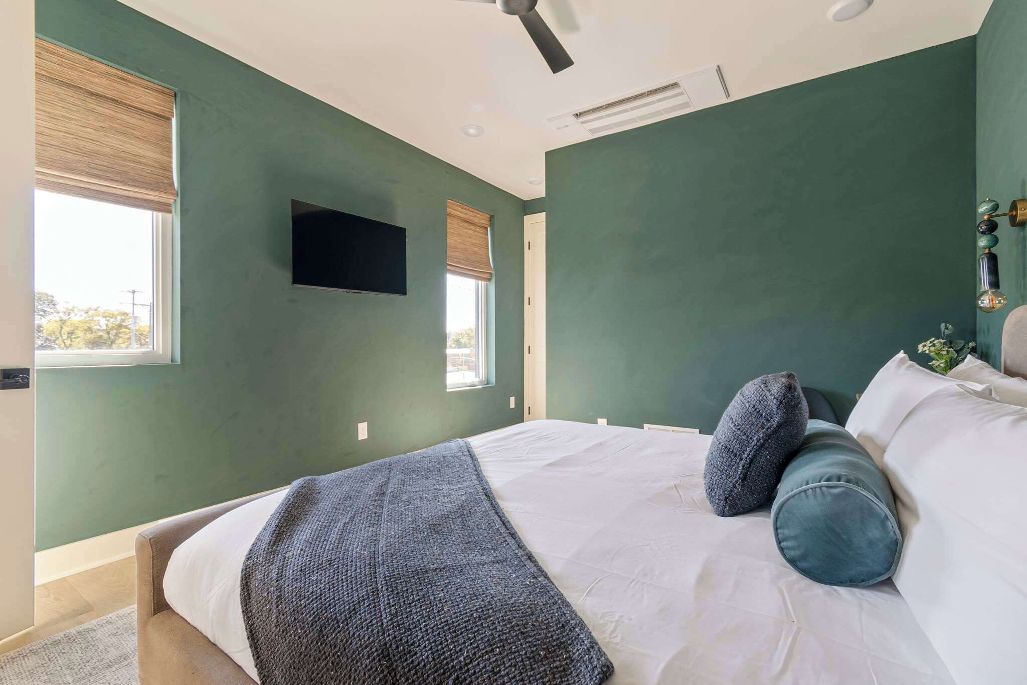 A bedroom with a bed, a ceiling fan, a flat-screen TV, and two windows with blinds.