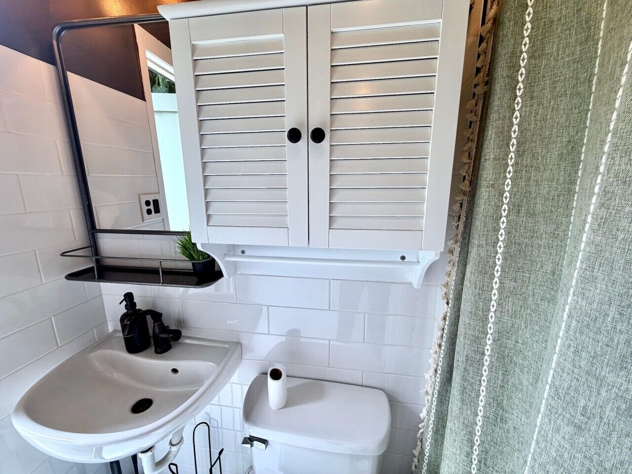 A bathroom with a white sink, toilet, and a mirror above the sink. There is a shelf with a plant and a black bottle.
