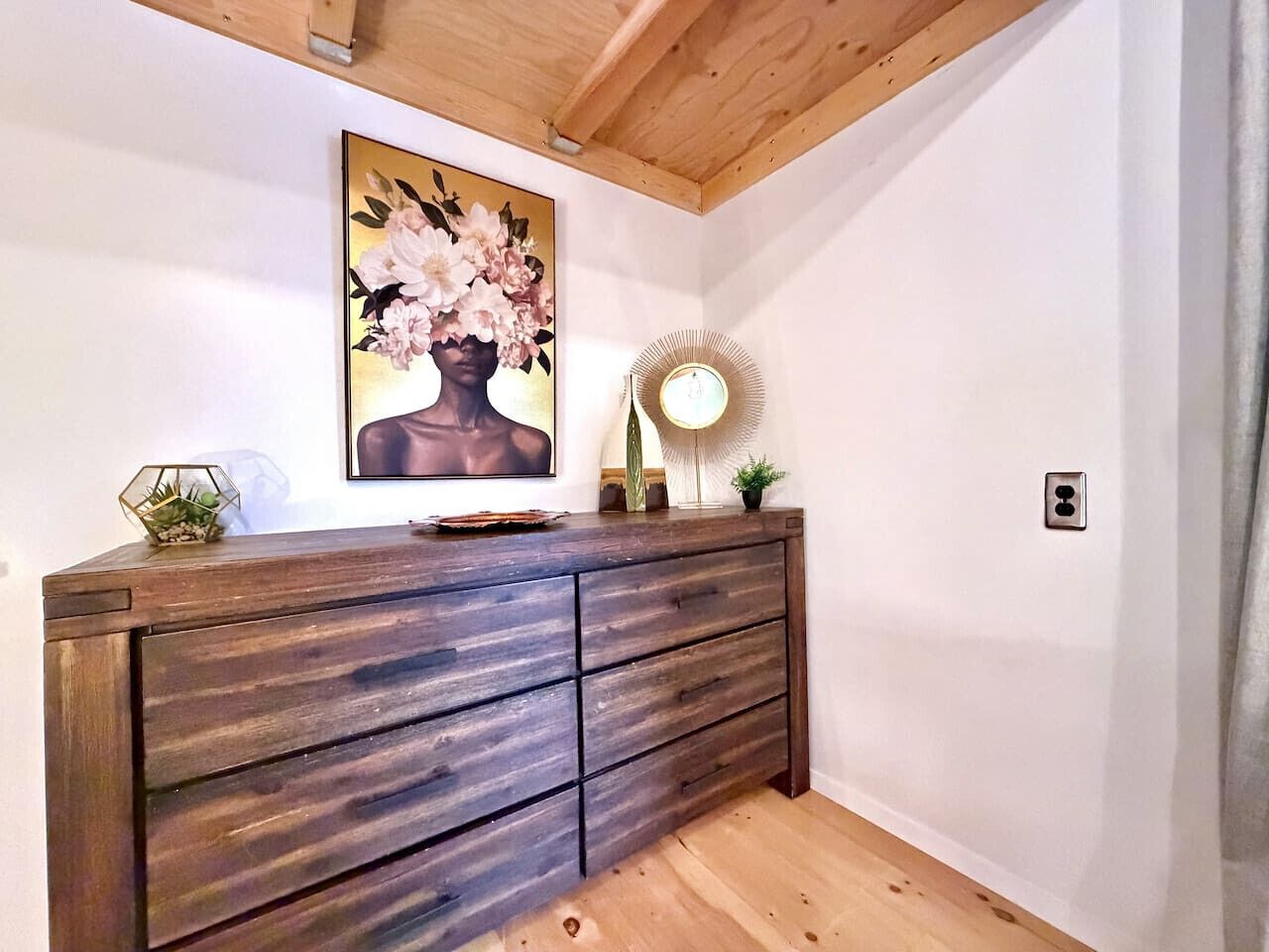 Un comò di legno con diversi cassetti, un quadro floreale incorniciato sopra, un piccolo tavolino con una lampada e un portavaso geometrico sopra.
