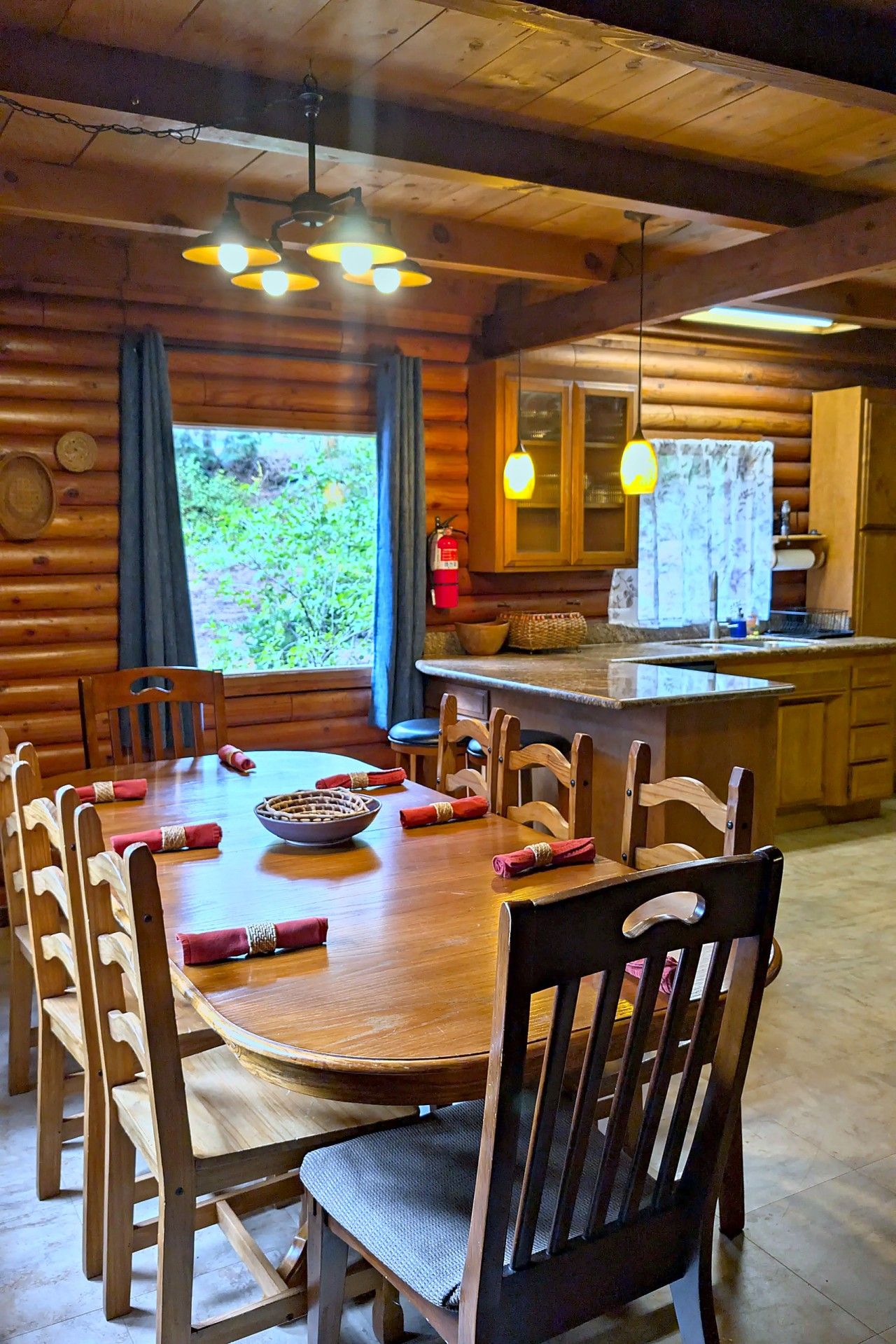 Interior de una cabaña de madera con una mesa de comedor para seis personas, una zona de cocina y un gran ventanal con cortinas.