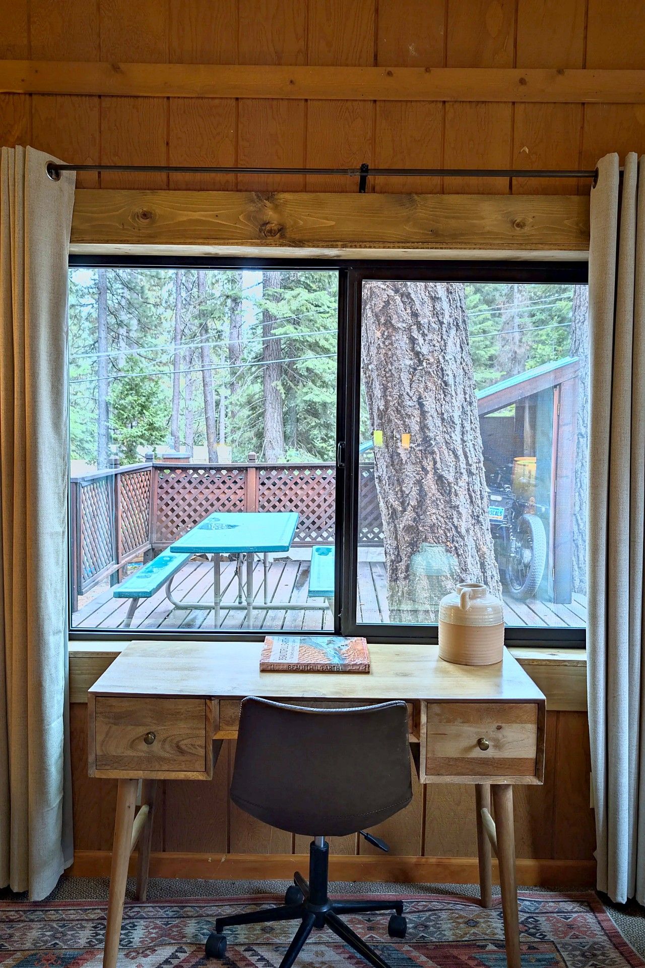 A wooden desk with a chair and a window overlooking a deck with a picnic table and a tree.