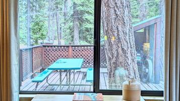 A wooden desk with a chair and a window overlooking a deck with a picnic table and a tree.
