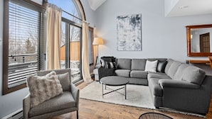 En moderne stue med en grå hjørnesofa, en mindre lænestol og et sofabord. Der er et stort vindue med persienner, en gulvlampe og et abstrakt kunstværk på væggen.
