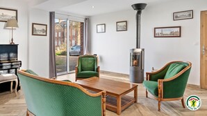 A modern living room with a piano, green upholstered chairs, a wooden coffee table, and a wood-burning stove. - Gîtes de France® Le cottage du parc (ROCQUEFORT)