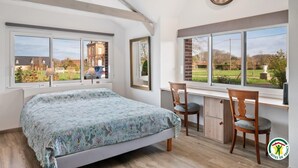 A bedroom with a bed, two chairs, a desk, and large windows offering a view of a garden and buildings. - Gîtes de France® Le cottage du parc (ROCQUEFORT)