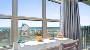 Ein Zimmer mit Schreibtisch, Blumentopf, Lampe, einer Uhr und Blick auf den Strand.