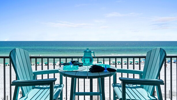 Un balcon avec deux chaises et une table, donnant sur une plage parsemée de parasols et où se trouvent des gens.