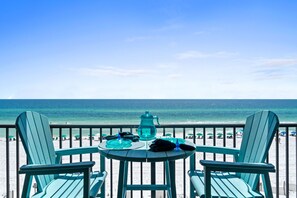 Un balcon avec deux chaises et une table, donnant sur une plage parsemée de parasols et où se trouvent des gens.