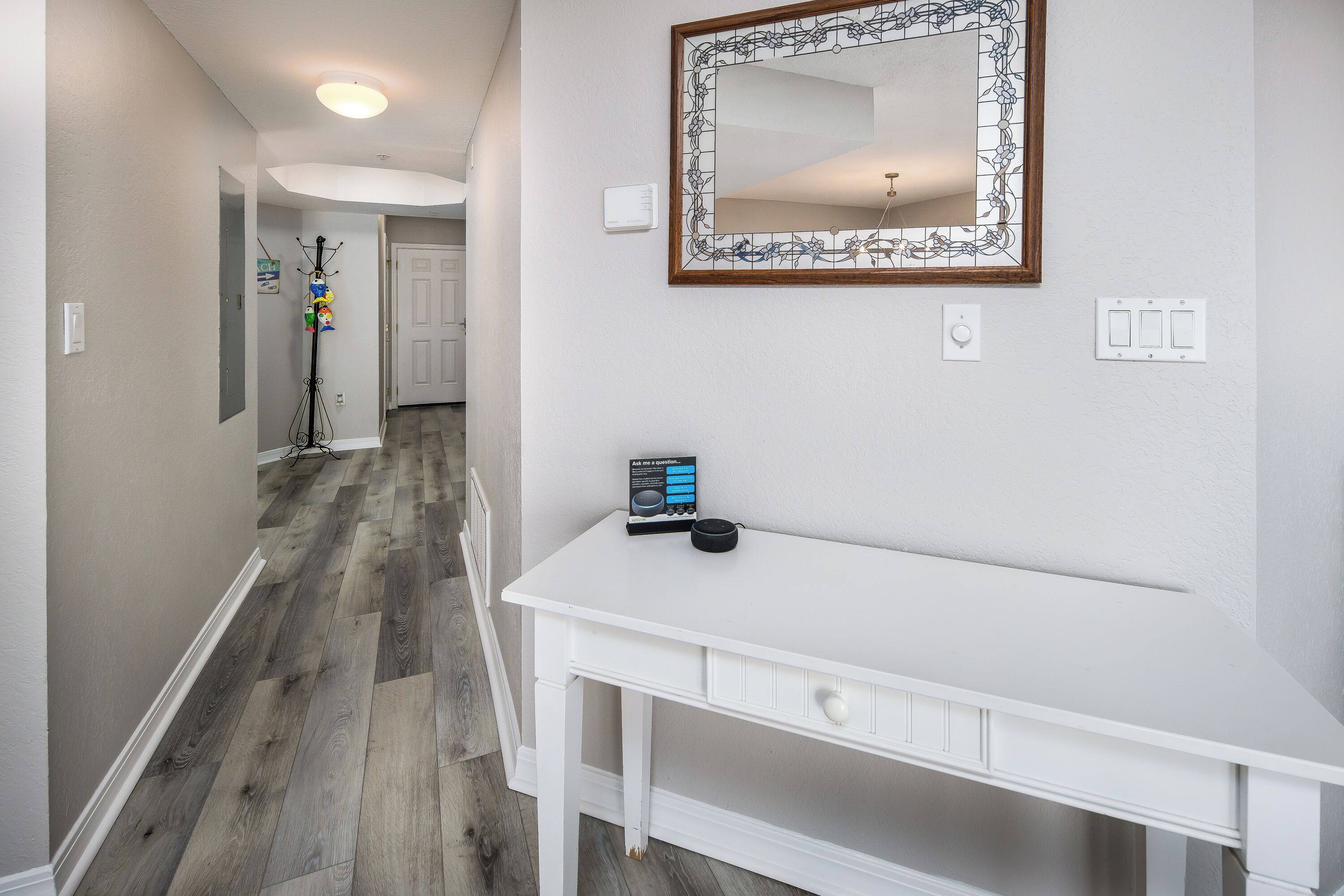 A hallway with a white desk, a mirror with a floral frame, and a door at the end.