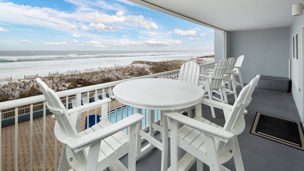 Un balcone con tavolo e sedie bianche, affaccia su una spiaggia con una passerella in legno e vista sull'oceano.