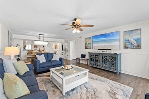 A living room with a blue sofa, a glass coffee table, a ceiling fan, a television, and artwork on the walls. - Surf Dweller 508 - Beachfront Boho (Fort Walton Beach)