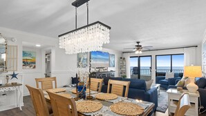 A dining area with a wooden table, chairs, and a chandelier. There is a living room with a blue sofa and a view of the ocean. - Surf Dweller 508 - Beachfront Boho (Fort Walton Beach)