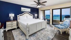 A bedroom with a large bed, a ceiling fan, a nightstand, a chair, and a balcony with a view of the ocean. - Surf Dweller 508 - Beachfront Boho (Fort Walton Beach)