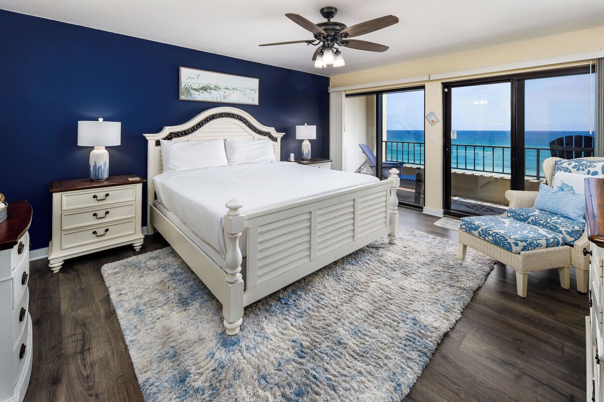 A bedroom with a large bed, a ceiling fan, a nightstand, a chair, and a balcony with a view of the ocean.