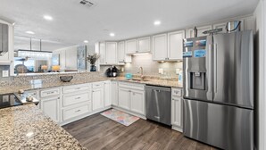 A modern kitchen with white cabinets, stainless steel appliances, granite countertops, and a wooden floor. - Surf Dweller 508 - Beachfront Boho (Fort Walton Beach)