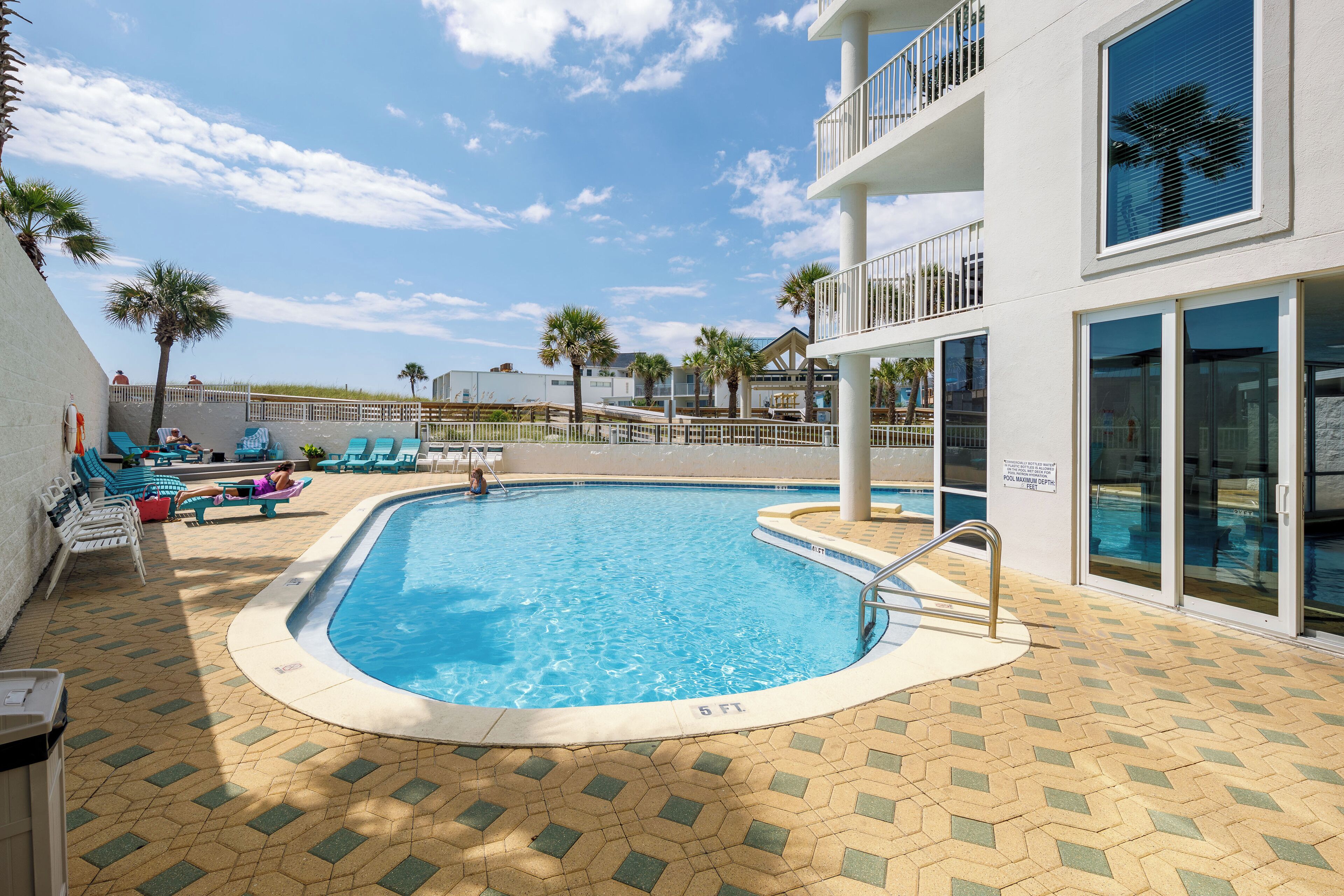 A swimming pool with lounge chairs and palm trees, surrounded by a building with balconies and large windows.