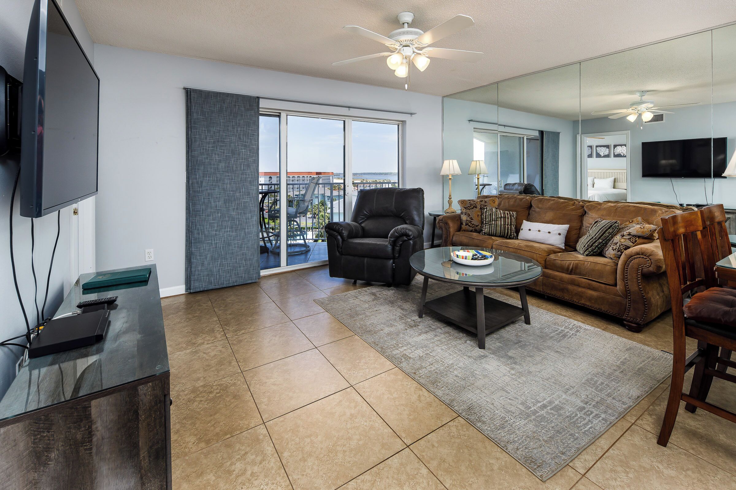 A spacious living room with a large sliding glass door leading to a balcony, a leather armchair, a brown sofa, and a flat-screen TV mounted on the wall.