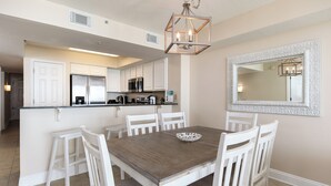 A dining area with a wooden table and white chairs, a kitchen with white cabinets and stainless steel appliances, and a decorative mirror on the wall. - Sea Dunes 402 - Sunny Dolphin (Fort Walton Beach)