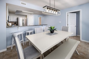 A modern kitchen with white cabinets, a stainless steel refrigerator, and a dining area with a white table and chairs. - Gulf Dunes 304- Saltwater Solace (Fort Walton Beach)