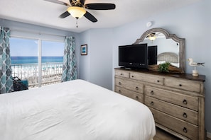 A bedroom with a large bed, a dresser, a television, a ceiling fan, and a view of the beach. - Gulf Dunes 304- Saltwater Solace (Fort Walton Beach)