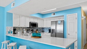 A modern kitchen with white cabinets, a central island with bar stools, a stainless steel refrigerator, and a microwave. - Gulf Dunes 210 - Coastal Charm (Fort Walton Beach)