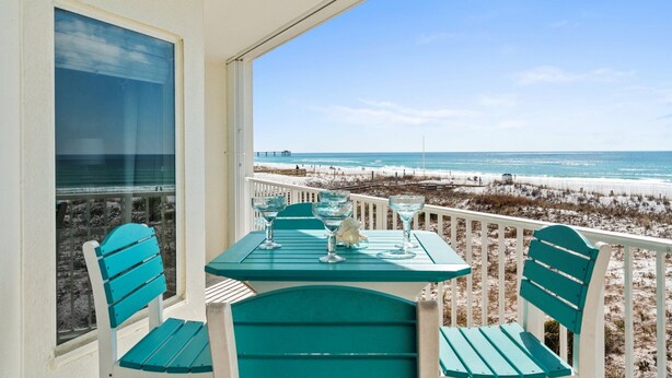 A balcony with a table and chairs, overlooking a beach with a fence and a pier in the distance. - Gulf Dunes 210 - Coastal Charm (Fort Walton Beach)