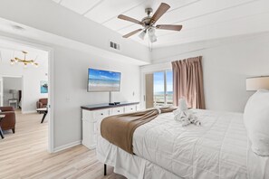 A modern hotel room with a large bed, a ceiling fan, a flat-screen TV, and a view of the ocean through the window. - Coconut Beach House A (Fort Walton Beach)