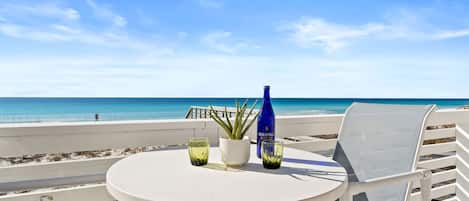 A round white table with a bottle and glasses on a balcony overlooking a beach.