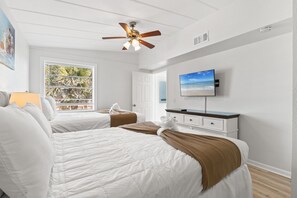 Camera d'albergo con due letti, una ventola a soffitto, una TV e una finestra vista palme.