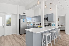 Una cucina moderna con mobili bianchi, un'isola grande con sgabelli da bar, elettrodomestici in acciaio inossidabile e illuminazione a sospensione.