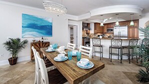 A dining area with a wooden table, white chairs, and blue glassware. There is a kitchen in the background with wooden cabinets and stainless steel appliances. - Bella Riva 108 - Golden Horizon (Fort Walton Beach)