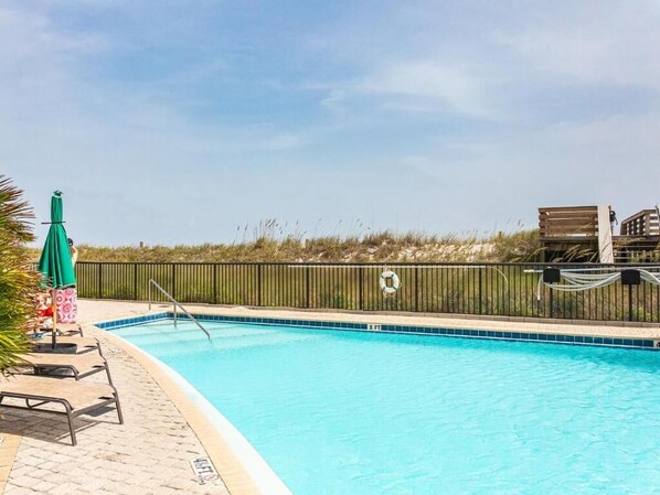 Une piscine avec un poste de surveillance pour le sauveteur et un parasol vert, entourée d’une clôture et d’une zone herbeuse.