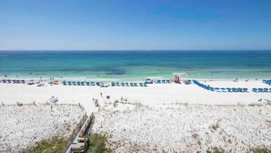 A beach with white sand and clear blue water. There are blue and red umbrellas and lounge chairs along the shore. - Sea Oats 704 - Coral Shores (Fort Walton Beach)