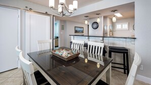 A dining area with a glass-top table, white chairs, a chandelier, and a kitchen in the background. - Sea Oats 704 - Coral Shores (Fort Walton Beach)