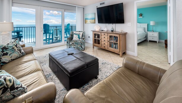 A spacious living room with a large TV, a comfortable sofa set, a wooden cabinet, and a balcony with a view of the sea. - Sea Oats 704 - Coral Shores (Fort Walton Beach)